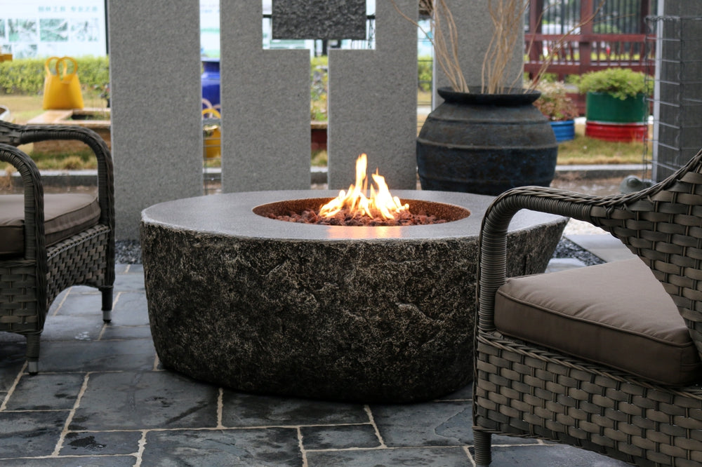Circular black stone firepit table with flames, surrounded by wicker chairs on stone patio