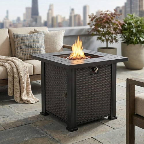 Black square steel fire pit table with wicker-style finish, lit flames, on stone patio near outdoor furniture