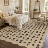 Beige jute rug with black floral pattern, scalloped edges, shown in bedroom on wooden floor