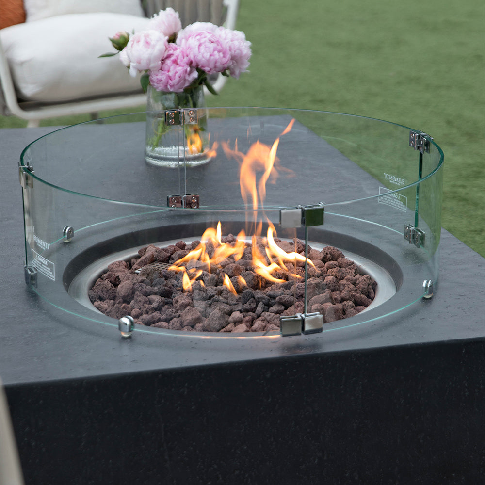 Dark grey fire pit table with glass surround and flames, outdoors on grass near patio furniture