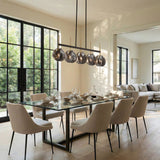Five-light linear chandelier with smoky gray shell glass shades, black frame, and gold accents, hanging over a dining table in a sunlit room