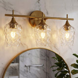 Brushed brass three-light vanity fixture with textured glass shades above a round mirror in a marble bathroom