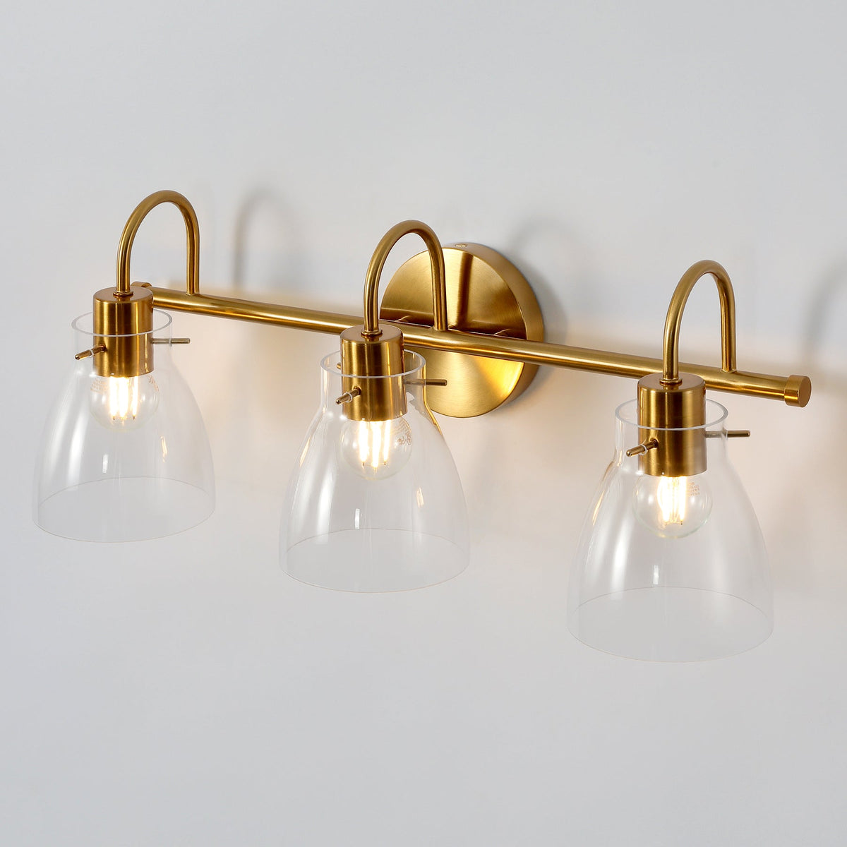 Three-light brushed brass vanity light with clear glass shades, shown from front angle