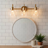 Three-light brushed brass vanity light with clear glass shades above a round mirror in a bathroom