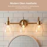 Three-light brushed brass vanity light with clear glass shades mounted above a mirror in a tiled bathroom