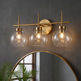 Brushed brass three-light vanity light with clear globe shades mounted above a round mirror