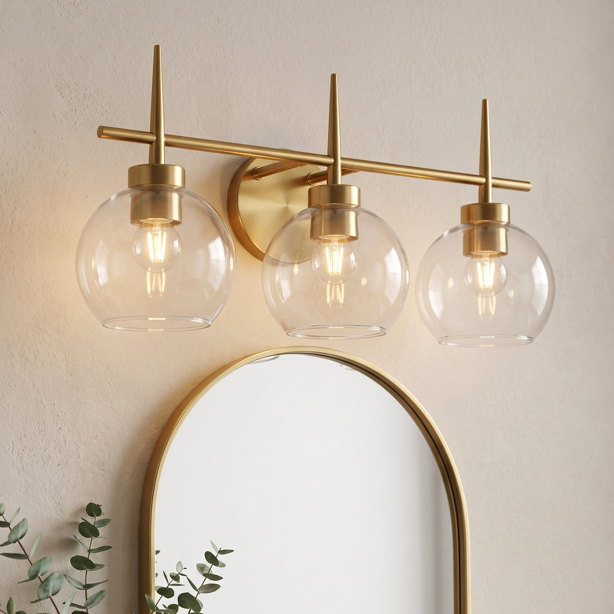 Three-light brushed brass vanity light with clear globe shades mounted above a mirror