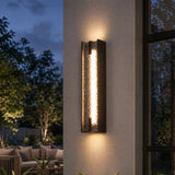 Black LED outdoor wall sconce with tall water ripple acrylic, mounted on wall near window at dusk