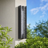 Black LED outdoor wall sconce with tall water ripple acrylic, mounted on wall beside greenery