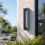 Black LED outdoor wall sconce with water ripple acrylic, mounted vertically on textured wall near patio