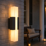 Black LED outdoor wall light with acrylic diffuser mounted on brick wall, glowing warmly on porch