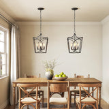 Two black metal geometric cage pendant lights hang over a wooden dining table in a room with beige walls and a window
